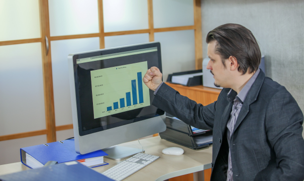 A man in a suit sits at a desk in an office, looking at a computer monitor displaying a bar chart with increasing values. He gestures towards the screen, appearing pleased. Office supplies and binders are seen on the desk.