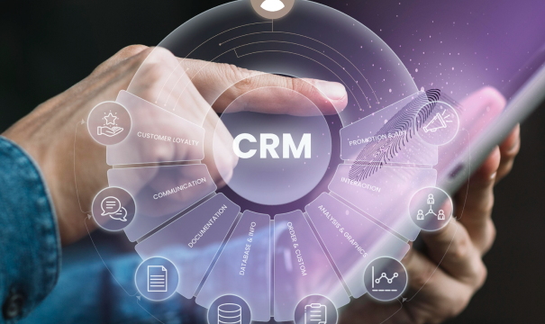 Close-up of hands holding a tablet with a floating digital CRM interface. The interface includes icons and circular graphics representing aspects like customer loyalty, documentation, communication, database, sales analysis, and promotions.