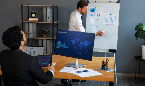 Two men in business attire are in a modern office. One is sitting at a desk, working on a computer with graphs on the screen. The other stands at a whiteboard, presenting charts.