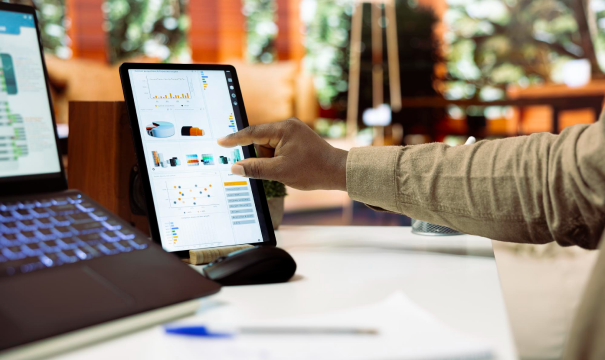 A person interacts with a tablet displaying charts and graphs on a desk. A laptop is partially visible on the left, and the background features a blurred office space with plants and furniture.