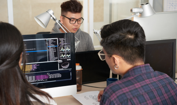 A group of individuals working at computer stations in an office setting. Two people in the foreground are focusing on a screen displaying code and graphs, while another person in the background is also working on a desktop computer. The scene is well-lit with desk lamps.