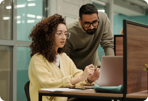 A woman with curly hair and glasses sits at a desk using a laptop, while a man with a beard and glasses stands beside her, leaning in to look at the screen. They appear to be working together in an office setting.