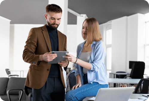 A man in a brown blazer and a woman in a light blue shirt are looking at a tablet together in a modern office setting. The woman is sitting on a table, and the man is standing beside her. Both appear focused on the tablet screen.