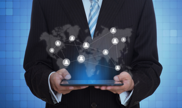 A person in a suit and tie is holding a tablet. On the tablet screen, there is an illustration of a world map with interconnected user icons, symbolizing global connectivity. The background consists of a gradient blue grid pattern.