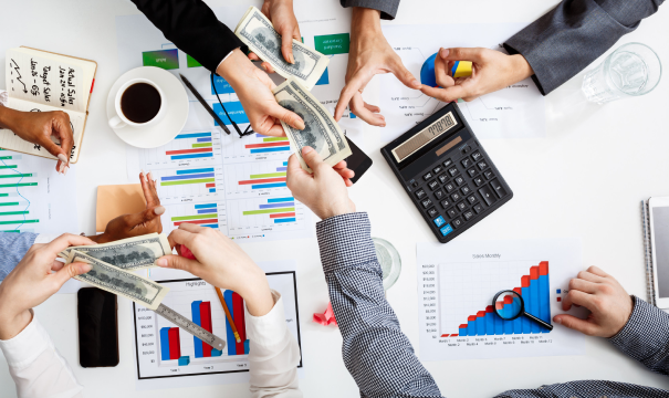 An overhead view of a table with business reports, a calculator, a magnifying glass, coffee, and hands exchanging cash. The charts display statistical graphs and data, indicating a business meeting or financial discussion.