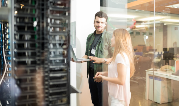 Two people stand in a modern office environment with large computer servers nearby. One person, holding a laptop, appears to be explaining something to the other. They are separated from the office space by a glass wall. The background shows open workspaces.