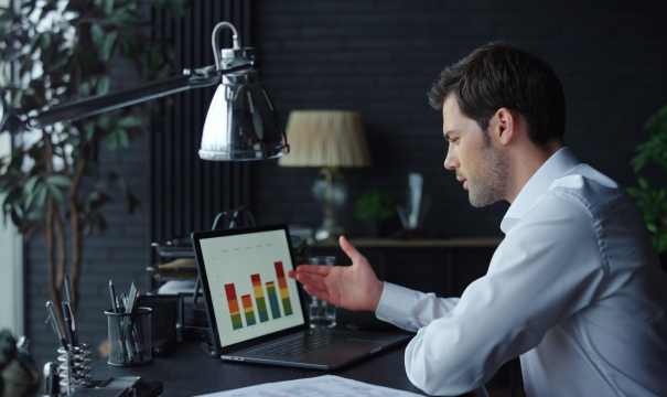 A man in a white shirt is seated at a desk, engaged in conversation via his laptop. The laptop screen displays colorful bar charts. The setting appears to be an office with dark walls, a lamp, and plants in the background.