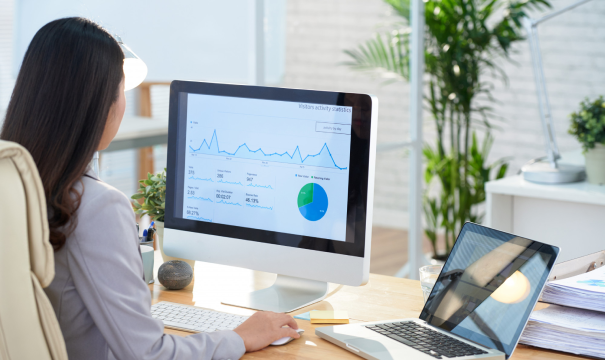 A person sitting at a desk looks at a desktop monitor displaying various charts and graphs, including a line graph and a pie chart. A laptop is open on the desk, and the workspace is adorned with plants and office supplies. Natural light illuminates the scene.