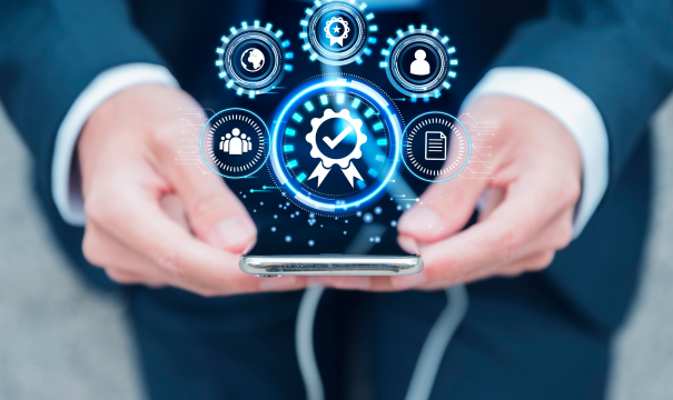 A person in a suit holds a smartphone. Over the phone, digital icons, including a globe, gears, and a checkmark with a ribbon, are displayed in a futuristic, glowing style, suggesting technology-driven concepts like certification, connectivity, and data management.