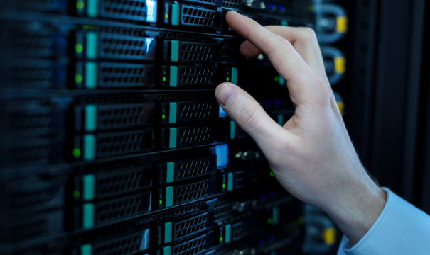 A person's hand is adjusting a piece of hardware on a rack-mounted server in a data center. The server has multiple slots with indicator lights, some glowing green. The environment is dimly lit, highlighting the server's intricate design and components.