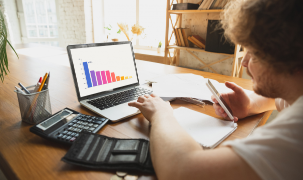 A person sitting at a wooden desk, analyzing a bar chart on a laptop screen. They hold a smartphone in one hand and point at the laptop screen with the other. The desk is cluttered with a calculator, notebook, pen holder, wallet, and some coins. A bookshelf is in the background.