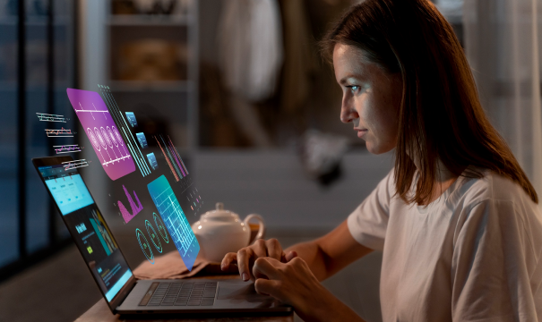 A woman in a white T-shirt is seated at a desk, typing on a laptop. Futuristic, holographic data charts and graphs are displayed above her laptop screen, highlighting data analysis. A teapot and a cup are on the table beside her. The background is blurred and modern.