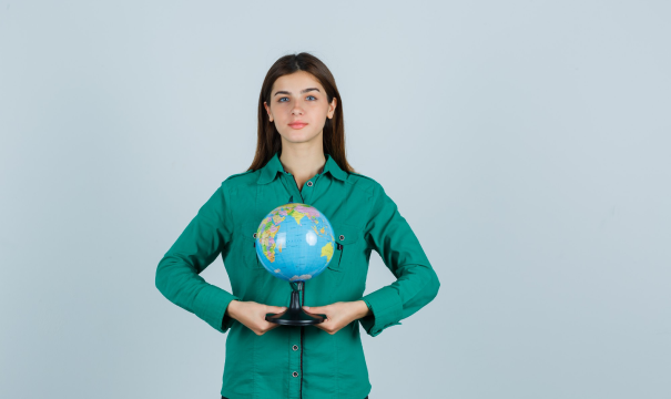 A woman with long brown hair, wearing a green shirt, holds a small globe in both hands against a plain, light grey background. She stands facing the camera with a neutral expression.