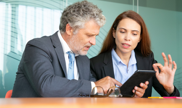 Two business professionals are sitting at a table in a modern office. The man with gray hair and beard is in a suit and tie, holding a tablet, while the woman with long brown hair in a blazer is gesturing and explaining something. A pair of eyeglasses lies on the table.