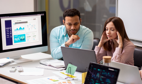 Two individuals are focused on a laptop in an office setting. A large monitor displays charts and graphs, while a tablet shows additional data analytics. Papers, notebooks, and other office supplies are scattered on the table around them. 