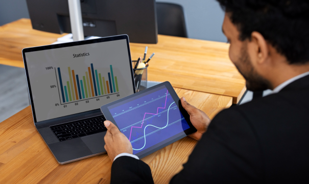 A person in business attire is seated at a desk, looking at a tablet displaying line and bar charts. A laptop next to them shows a bar graph titled 