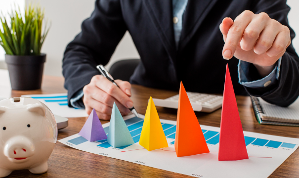 A person in business attire touches a red cone-shaped figure on a financial chart with colorful 3D paper pyramids. A piggy bank, pen, papers, calculator, and potted plant are on the wood desk. The scene suggests financial analysis or planning.