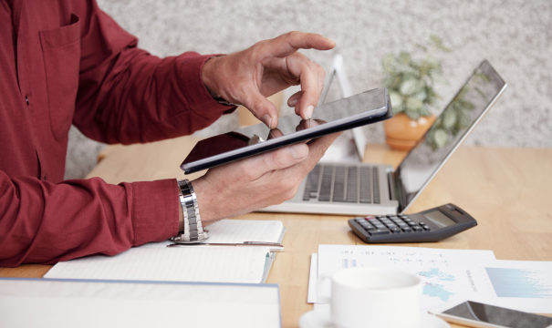 A person in a red shirt using a tablet at a cluttered desk with a laptop, papers, a calculator, a notepad, a pen, a coffee cup, and a small plant in the background. The workspace appears busy and organized for multitasking.