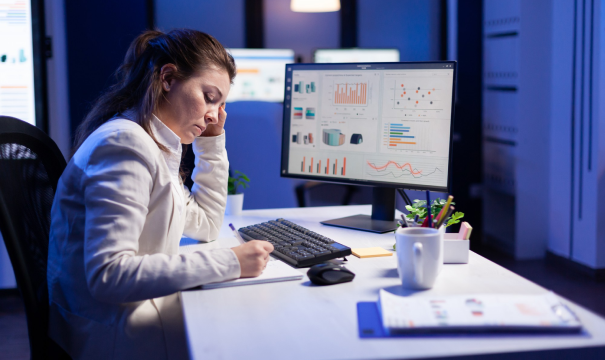 A woman in a white blazer sits at a desk, concentrating on writing in a notebook and is leaning on her hand. A computer monitor on the desk displays various charts and graphs. The background is a dimly lit office space.