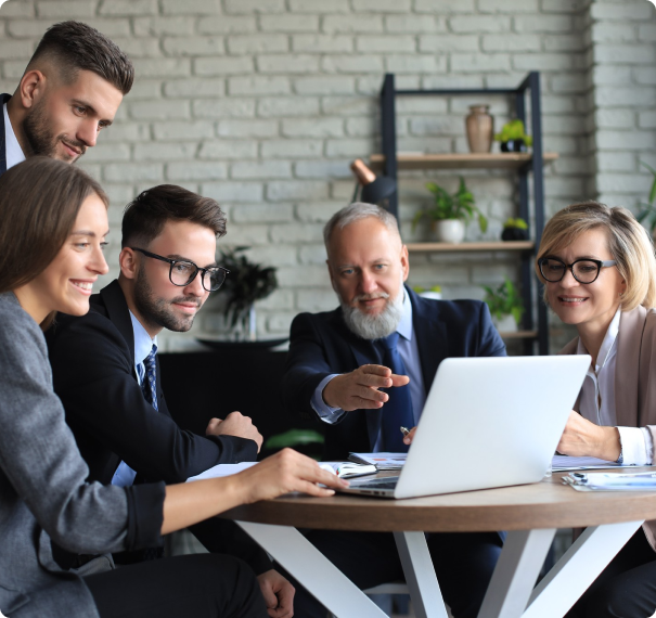 A group of five business professionals, three men and two women, are gathered around a table looking at a laptop screen. They are all dressed in business attire and appear to be engaged in a discussion. The setting is a modern office with shelves and plants in the background.