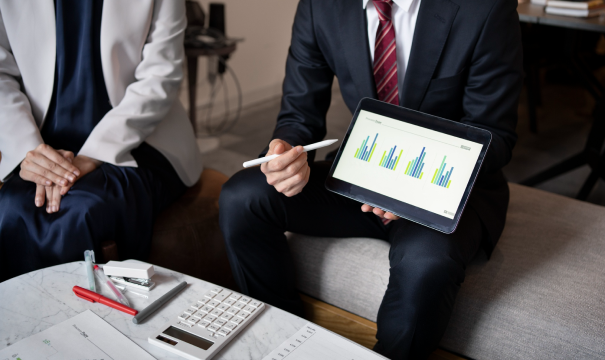 Two business professionals sit side by side, one holding a tablet displaying bar charts. Documents, a calculator, and office supplies are on the table in front of them, suggesting a financial meeting or presentation. They are engaged in a discussion.