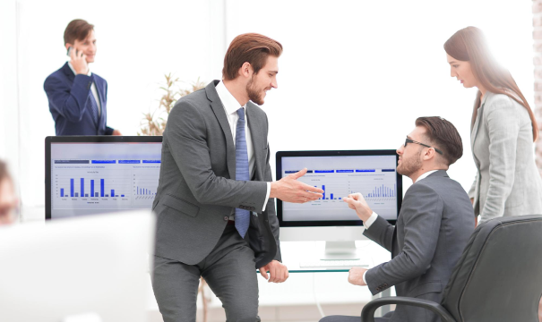 Four business professionals in an office setting. One person is on the phone, two people in suits are shaking hands, and another person is observing while seated at a desk. Computer monitors in the background display graphs and charts.