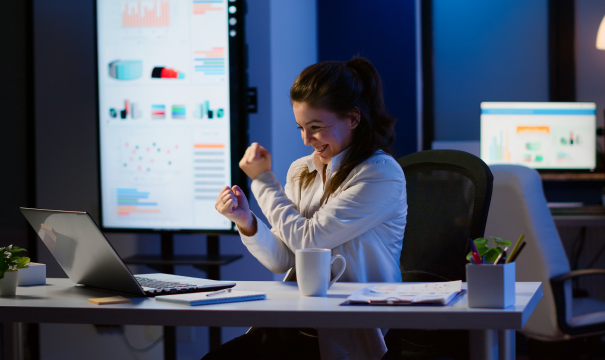 A woman sitting at a desk is celebrating while looking at her laptop. The office is dimly lit, and there are charts and graphs displayed on large screens in the background. She has a coffee mug and office supplies on her desk.