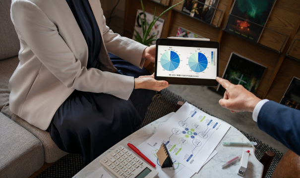 Two individuals in business attire are in a meeting. One person holds a tablet displaying charts with blue and green segments while the other points at it. On the table are papers with diagrams, a smartphone, calculator, markers, and a laptop.