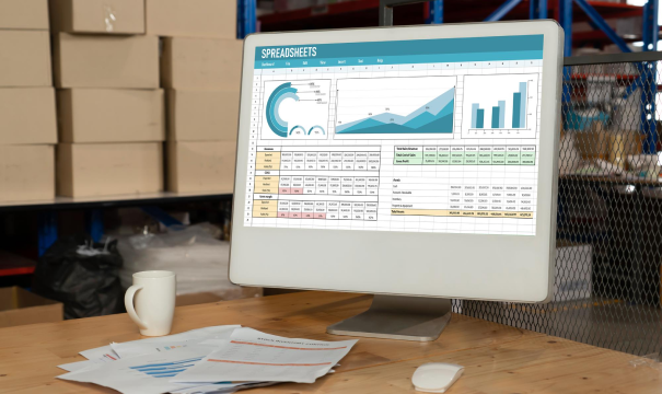 A computer monitor displaying a detailed spreadsheet with various charts and graphs. The setup is on a wooden desk with paperwork, a white coffee mug, and a computer mouse nearby. The background shows shelves of boxes in a storage or warehouse setting.