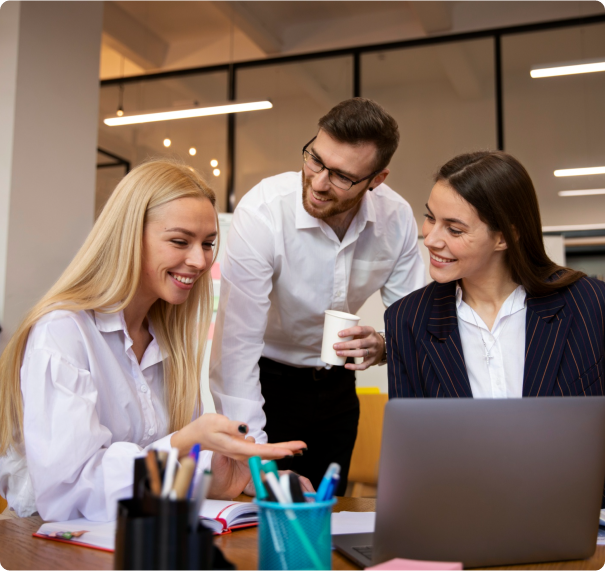 Three colleagues in a bright office are gathered around a laptop. A woman with long blonde hair is smiling and gesturing, a bearded man is holding a coffee cup and leaning forward, and another woman with long dark hair is smiling, all wearing business attire.