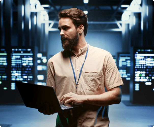 A person with a beard and wearing a lanyard is working on a laptop in a dimly lit, high-tech server room. The background features rows of server racks with glowing lights, creating an advanced technological atmosphere.