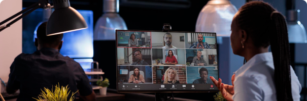 A woman gestures towards a computer monitor displaying a video conference with ten participants. She stands in a modern office with a desk and lamp visible. Another person is seated at a desk in the background, working on a computer. Potted plants are on the desks.