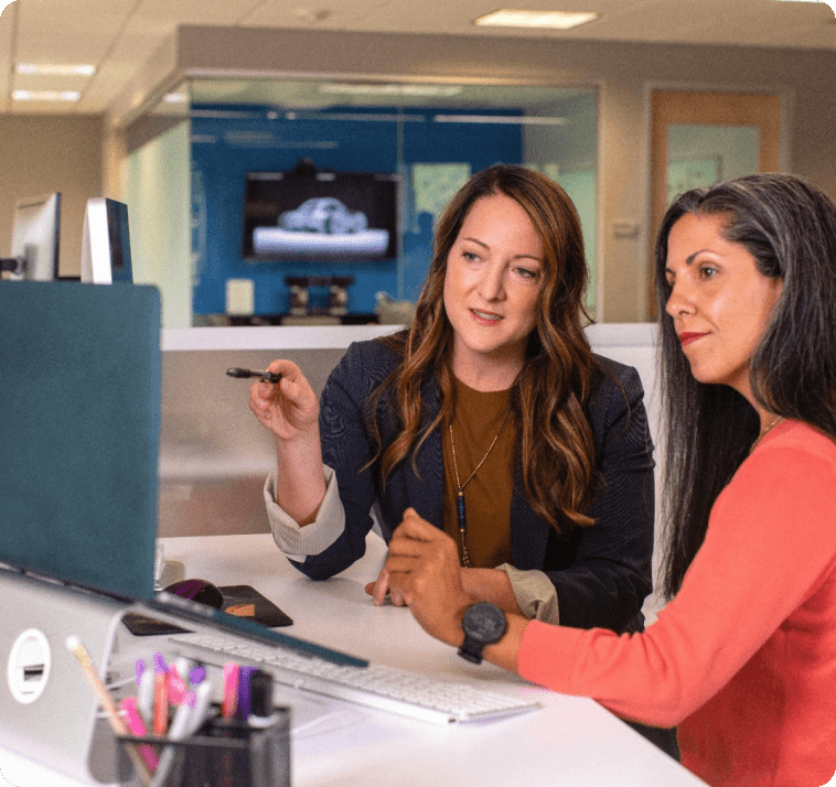 Two women sitting at a desk in a modern office, looking intently at a computer screen. One woman, with wavy hair, is pointing at the screen with a pen, while the other, with straight hair, listens attentively. Office supplies are visible on the desk.